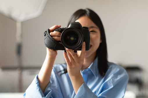 Portrait Of Happy Photographer Lady Taking Photo Holding Camera Near Face And Smiling Standing In Modern Studio Indoors. Professional Photography Art And Career, Creative Professions Concept