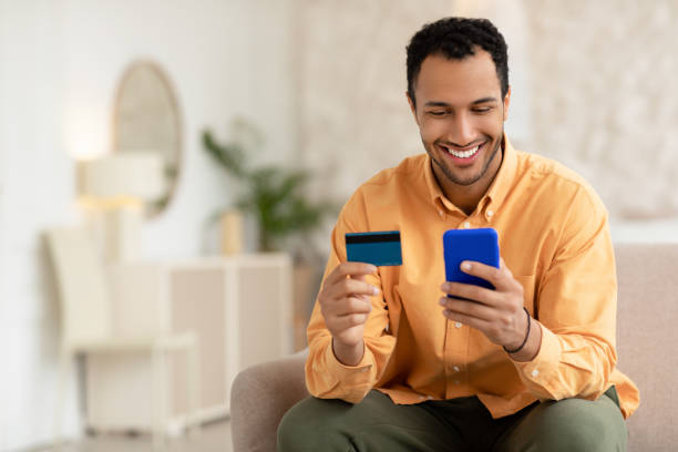 istock Smiling young guy using cellphone and credit card at home 1471032719