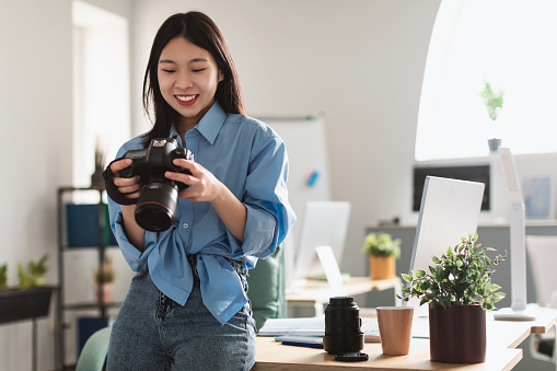 Portrait Of Smiling Asian Female Photographer Holding Camera Working With Photos, Reviewing Looking Through Taken Pictures At Office, Sitting Leaning On Table Desk. Art And Creative Profession Concept
