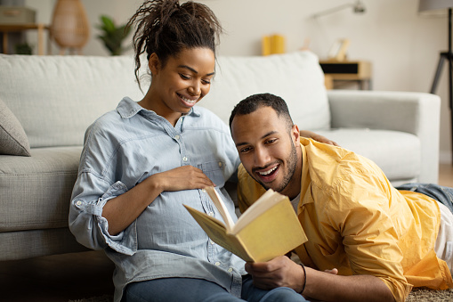 Happy Pregnancy. Loving African American Couple Reading Book Together Expecting Baby Sitting On Sofa At Home On Weekend. Family Lifestyle, Love And Childbirth Concept