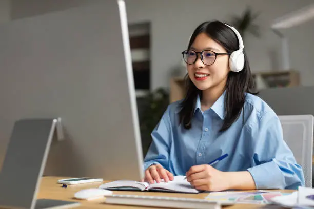 Distance Education. Portrait of smiling Asian woman sitting at desk, using pc and writing in notebook, taking notes, watching tutorial, lecture or webinar, studying online at home looking at screen