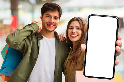 Shopping App. Cheerful Buyers Couple Showing Smartphone With Big Empty Screen To Camera Recommending Mobile Banking Application Standing In Modern Mall. Selective Focus