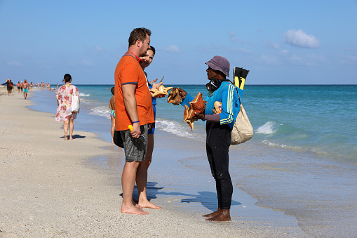 Varadero, Cuba - February 2023: Black man offers dried starfish and seashells to tourists on sea beach. Souvenir seller on tropical resort