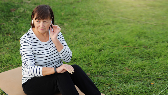 Middle aged woman in casual sportswear and wireless headphones choosing music for workout, sitting on fitness mat and using application on smart watch. Healthy active lifestyle and sport concept.