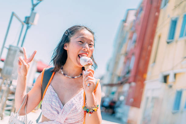 Portrait of a Cheerful Chinese Asian Tourist Young Woman Licking Sweet Italian Gelato Ice Cream while Visiting Venice Italy on Holiday in the Summer with Her Fingers Making a "Peace" or "Victory" Sign Portrait of a Cheerful Chinese Asian Tourist Young Woman Visiting Venice Italy on Holiday during the Hot Summer licking ice cream cone stock pictures, royalty-free photos & images