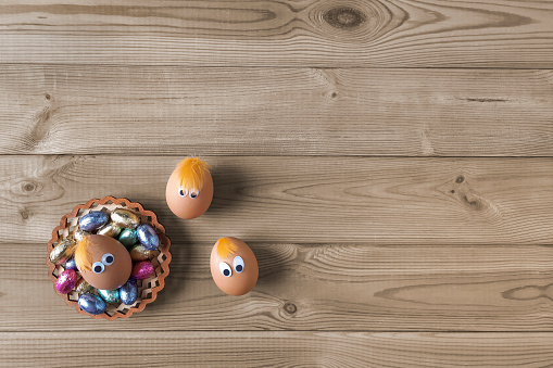 Three eggs with eyes and plate of chocolate Easter sweets on wooden table. Copy space. Top view.