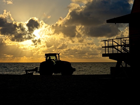 Beach cleanup as the sun rises over Miami's South Beach with lifeguard hut in silhouette. A sand grader or tractor grooms the beach as warm lit cumulus clouds shine over the Atlantic ocean