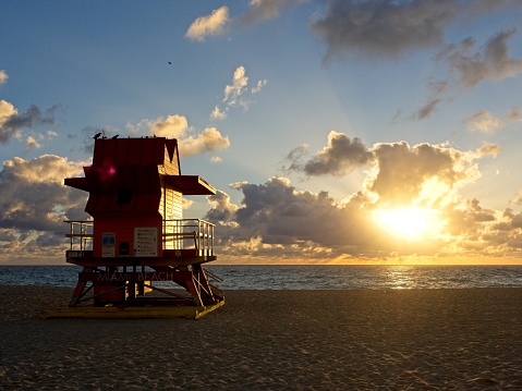 Sun rises over Miami's South Beach with lifeguard hut in silhouette. Blue sky with warm lit cumulus clouds over the Atlantic ocean and attendant corpuscular sun rays.