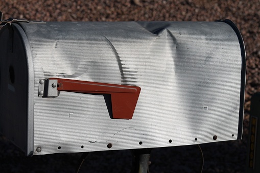 A silver metal mailbox with a large dent and a red flag.