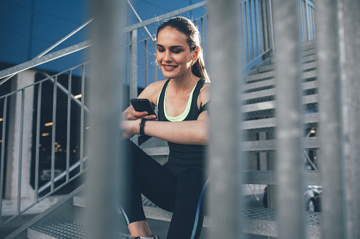 Fit female runner using smart watch and mobile phone to monitor her performance. Sportswoman checking her workout progress. Using smart technologies after workout outdoor in the city.