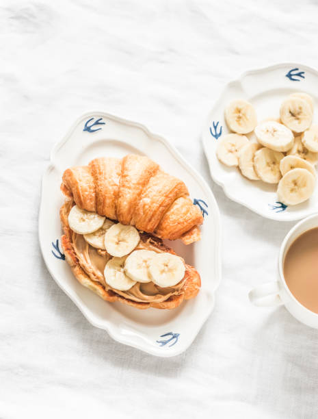 Cozy breakfast - croissant with peanut butter, banana and coffee with cream on a light background, top view