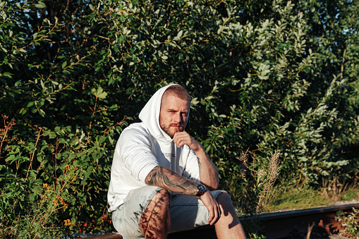 Bearded jewish man with tattoo in white jacket sitting on railroad, looking at camera. Male spending summertime in countryside outside. Lifestyle concept walk in fresh air nature. Copy ad text space