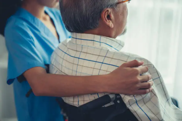 Rear view of a caregiver and her contented senior patient gazing out through the window. Elderly illness, nursing homes for the elderly, and pensioner life