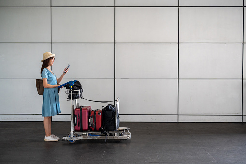 An Asian woman with a luggage cart using phone and waiting for a ride to pick her up at airport terminal.