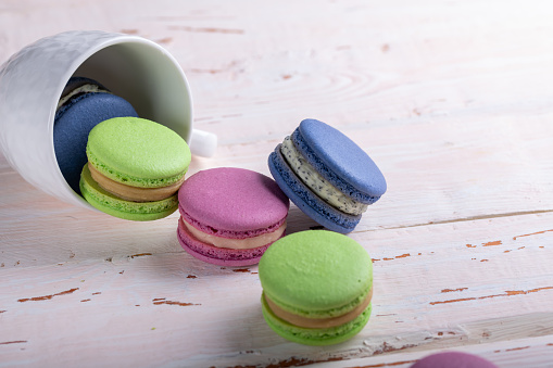 Colorful macaroons crumble from a white cup on a light wooden table, copyspace