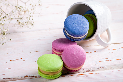 Colorful macaroons crumble from a white cup on a light wooden table, copyspace