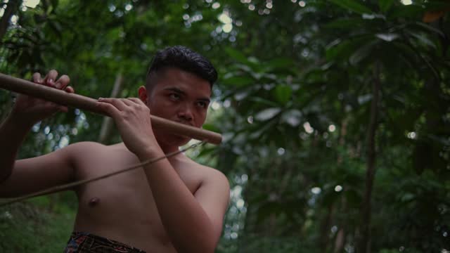an Asian boy is playing a bamboo flute very seriously in a forest early