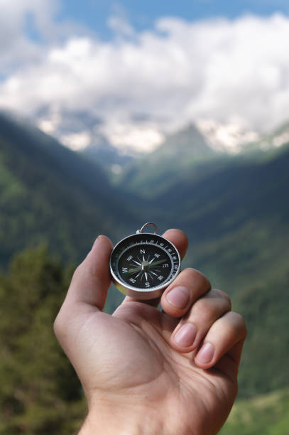 Magnetic compass in the palm of a male hand against the backdrop of a mountain range in the clouds in the summer outdoors, travel, first-person view stock photo