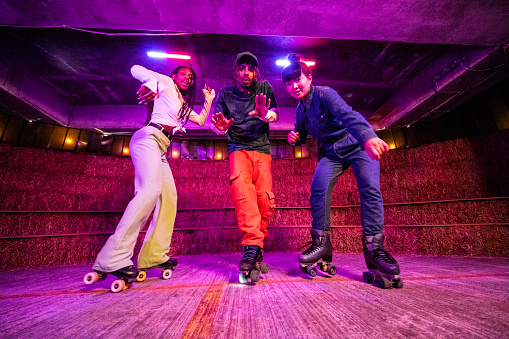 Low angle full length view of Black and Asian roller skaters in 20s and 30s wearing retro outfits, showing their dance moves, looking at camera.