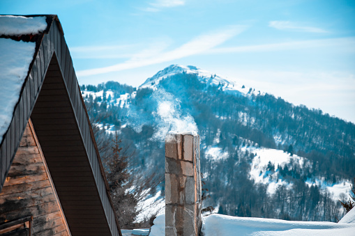 Smoke from the chimney on the roof of the house against blue snowcapped mountains. Copy space on blue sky