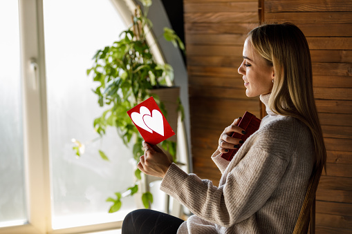 A charming woman sitting on a wooden chair, holding a gift and a greeting card for Valentine's Day, enjoying her reading.