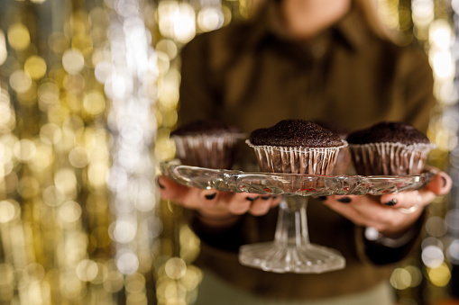 A woman showcasing a dessert tray filled with chocolate muffins, with a festive backdrop in the background.