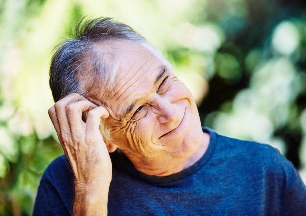 Man scratches his head as he thinks deeply about a choice or decision, as he stands outdoors among brightly lit foliage.