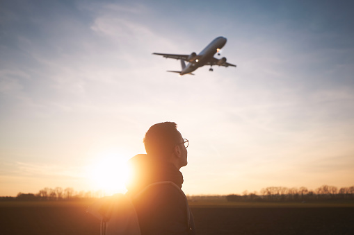 Man with backpack looking up to airplane landing at airport during beautiful sunset.