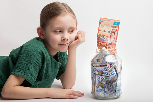 Kid with a large transparent jar with Kazakhstani tenge money on a white background.