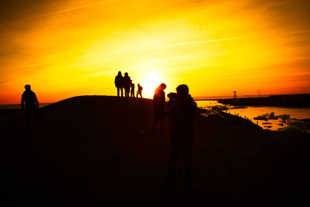 Mature adult female latin tourist standing in silhouette against a vibrant purple and orange sunset sky, embracing outdoor adventure and wellness on the sand dunes.She wears exercise pants, a sports jacket for the cold weather, and a scarf.