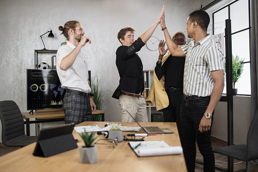 Diverse financial male traders giving high five to each other after productive cooperation at office. Competent brokers standing near desk with various modern technology and papers.