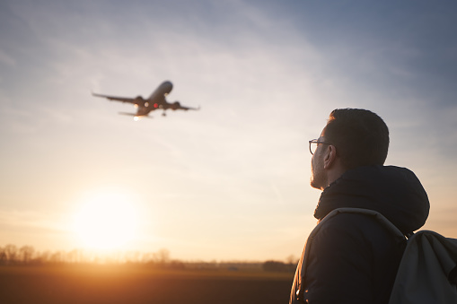 Man with backpack looking up to airplane landing at airport during beautiful sunset.