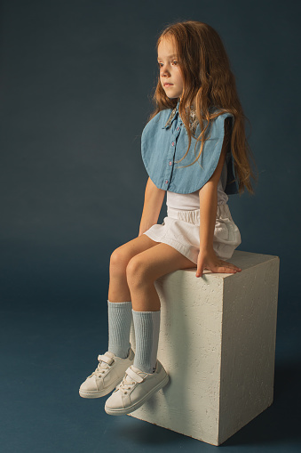 Dreaming, looking up. Portrait of little cute girl in casual clothes sitting on big box isolated on white studio background. Happy, joyful childhood, kids fashion, emotions, facial expression concept