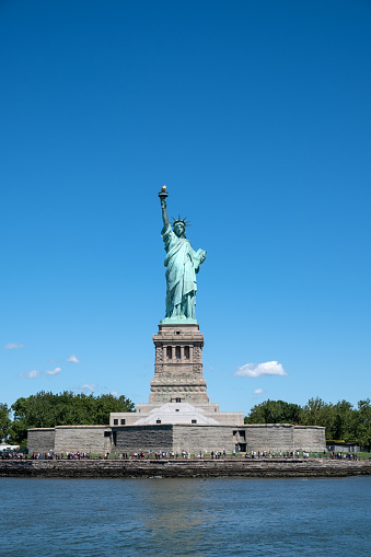 The iconic Statue of Liberty stands tall and proud at the mouth of the Hudson River against the backdrop of a sunny blue sky.
