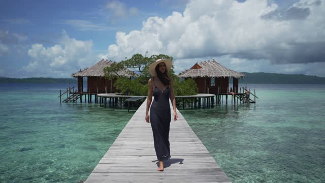 View of a young woman walking on a raised walkway above tropical waters with cabana cabins in the background