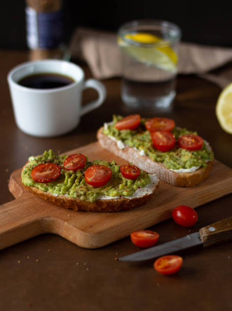 Toast with avocado and cherry tomatoes on a wooden board with a cup of coffee, a glass of water with lemon.