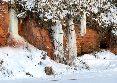 winter landscape with red sandstone cliffs and icefall on the bank of the river Salaca, the sun shines on the trees and the bank of the river, the ice covers the river, Sarkanas clifts, Latvia