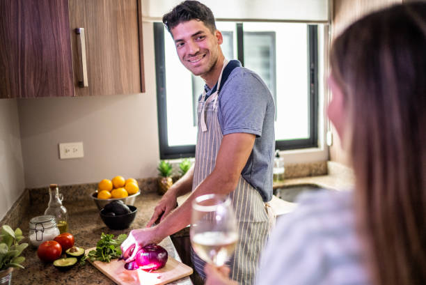 young man cutting vegetables and talking to his wife in the kitchen at home - routine foods and drinks clothing household equipment imagens e fotografias de stock