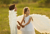 Two angels with white wings on green grass. Blonde girl in dress and brunette boy on summer sunset background