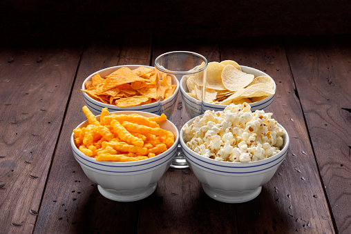 View of four bowls and empty glass in the center over rustic wood table. In the bowls the appetizers are : Cheese sticks, popcorn, fried potato chips and fried corn flakes with cheese. Ready appetizers to watch a movie at home with the family.