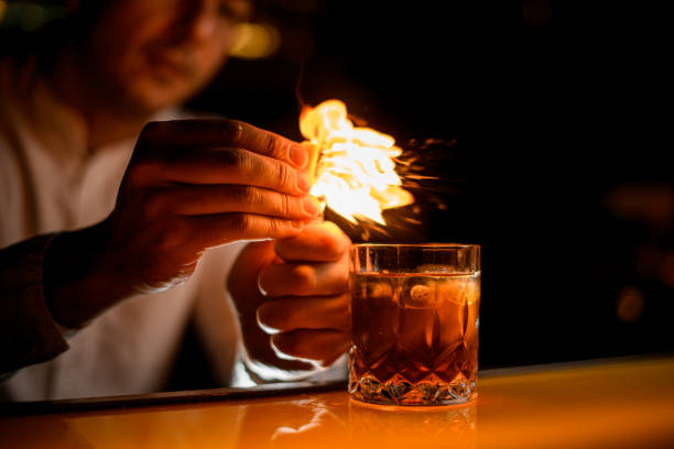 view on glass with drink on bar and male hand sprinkles by zest and sets it on fire - transparent holding glass focus on foreground imagens e fotografias de stock