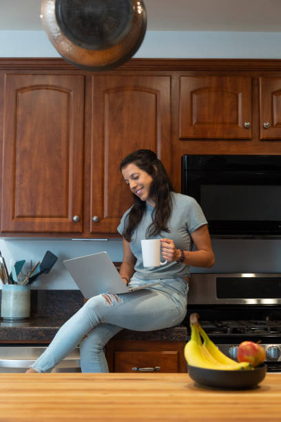 latynoska kobieta siedząca przy kuchennym blacie z laptopem i pijąca kawę - stereotypical housewife women domestic kitchen brown hair zdjęcia i obrazy z banku zdjęć
