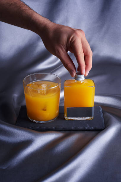 frasco de vidrio con bebida de naranja, en el fondo un vaso con hielo y frutas secas sobre un fondo gris de tela, sostiene a mano la tapa del frasco - restaurante sketch fotografías e imágenes de stock