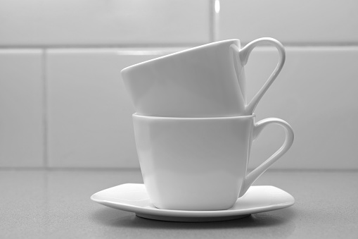 two white coffee mugs standing one on top of the other on a white saucer in black and white
