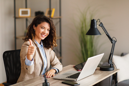 Portrait of a young business woman sitting at a desk in the office looking at the camera and giving a thumbs up