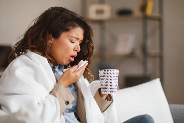 Mujer joven tosiendo usar papel pañuelo de papel sostiene una taza de té y está envuelto en una manta en casa - foto de stock