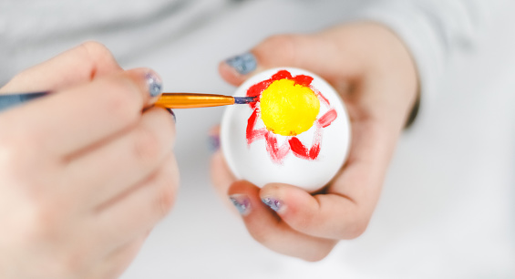 Hands of a caucasian girl painting an Easter egg with a brush with red and yellow acrylic paint at a marble table for diy preparation for the Easter holiday, close-up side view. The concept of crafts, needlework, artisanal, children art, diy, at home.