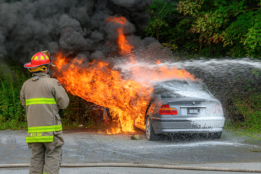 Saint John, NB, Canada - August 18, 2022: A parked car engulfed in flames at the side of a road. A stream of foam water fights the fire as a fireman watches. Trees in the background.