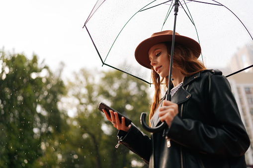Side view of serious young woman in hat using typing smartphone holding transparent umbrella standing in rainy autumn city waiting for taxi car. Concept of modern female lifestyle at autumn season.