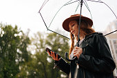 Side view of serious young woman in hat using typing smartphone holding transparent umbrella standing in rainy autumn city waiting for taxi car.
